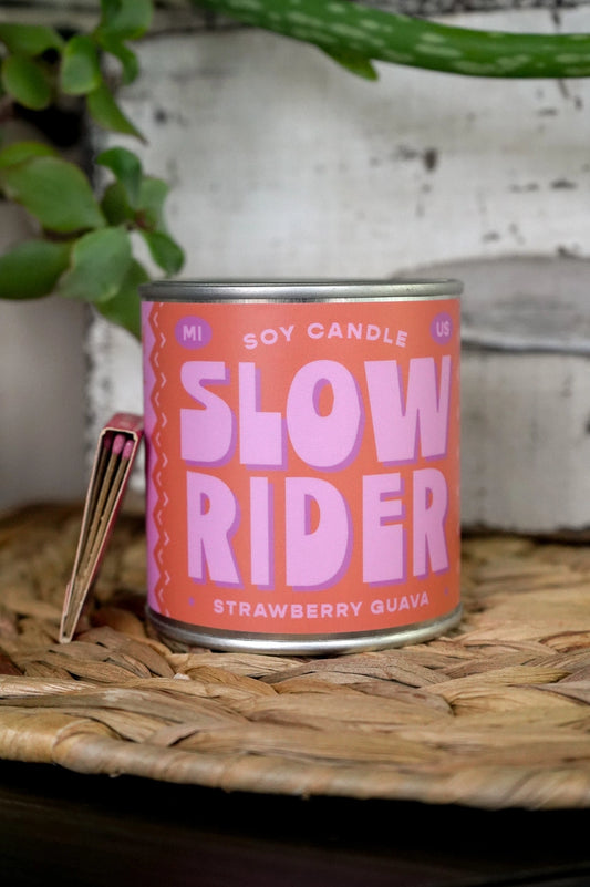 Candle tin labeled 'Slow Rider' on a woven surface with plants in the background