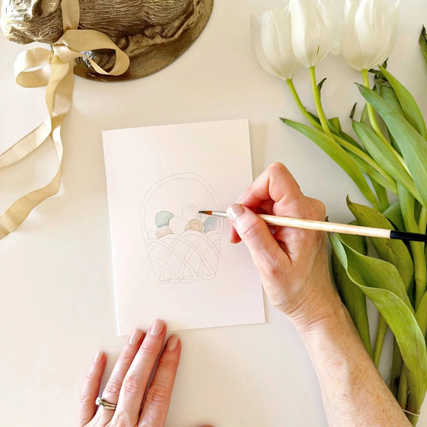 Person painting on a card with white tulips and ribbons in the background