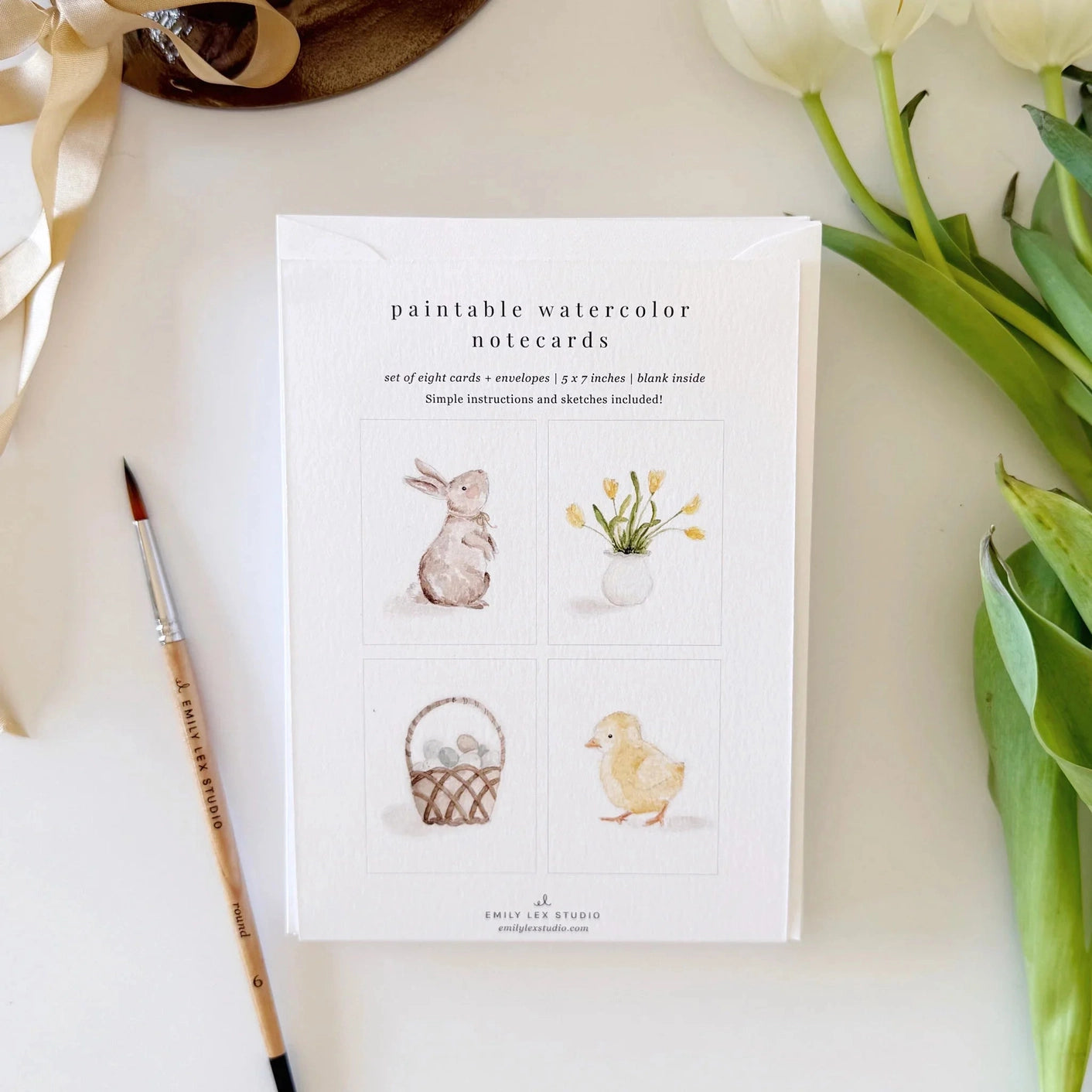 Set of paintable watercolor note cards with illustrations on a white surface with flowers and a brush.