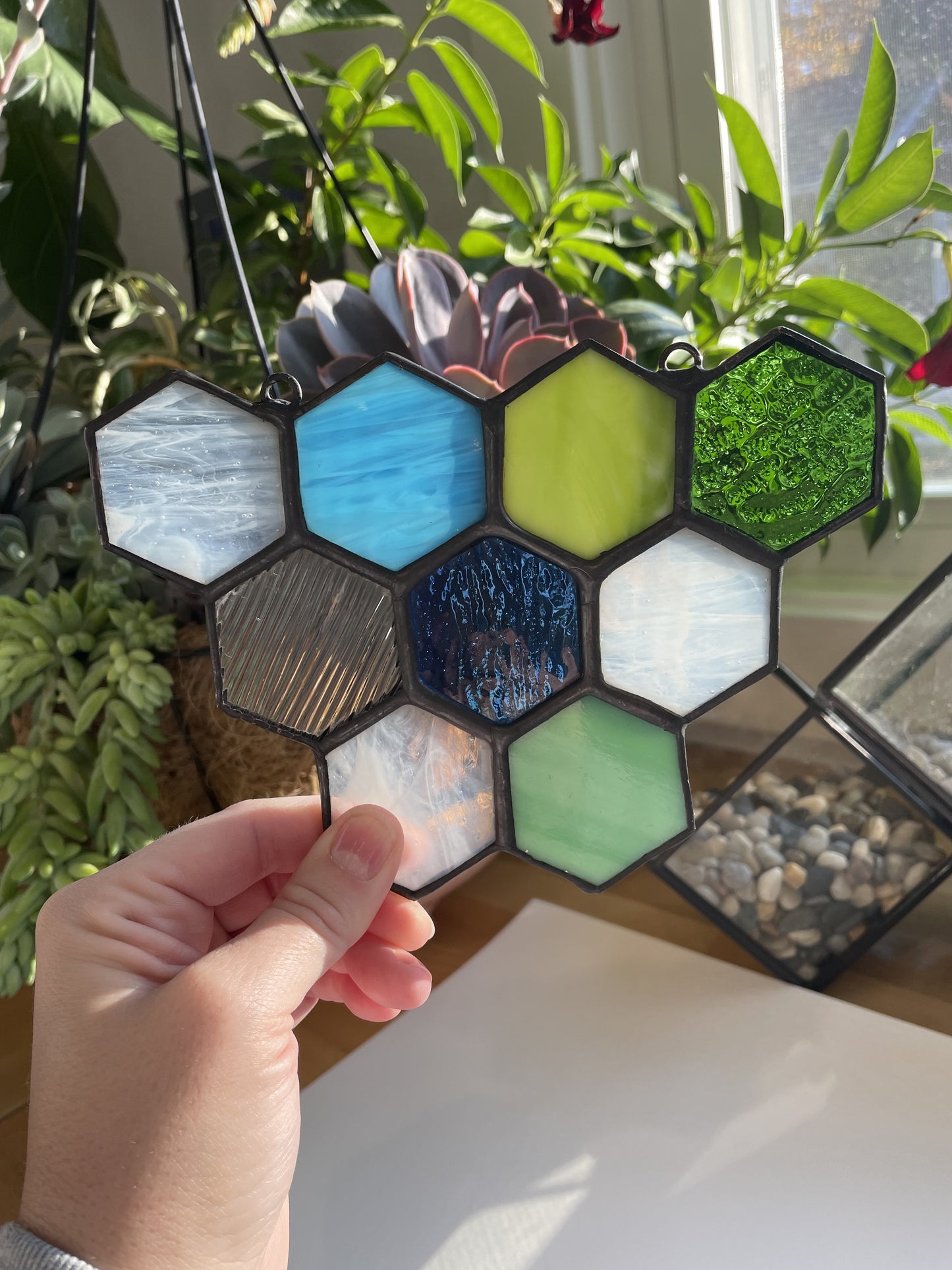 Hand holding a hexagonal stained glass panel with various colors and textures, surrounded by plants.