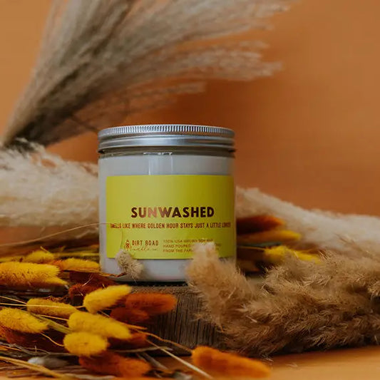 Candle jar labeled 'Sunwashed' surrounded by dried plants on a warm-toned background