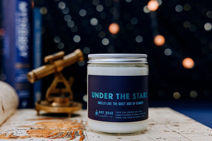 Candle jar labeled 'Under the Stars' on a table with decorative items and blurred lights in the background