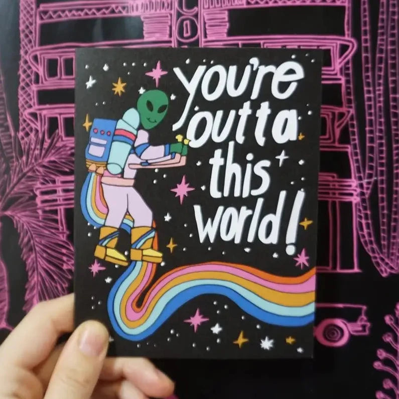 Card with alien design and text 'you're outta this world!' held against a pink and black patterned background.