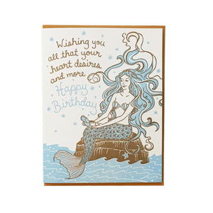White card with a blue and brown mermaid on a rock illustration. Gold text reads "wishing you all that your heart desires and more." Blue text reads "happy birthday"
