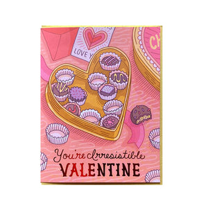 White card with pink background and a yellow heart-shaped box of chocolates. Some chocolates have been eaten.Shiny pink text reads "you're irresistible, valentine"