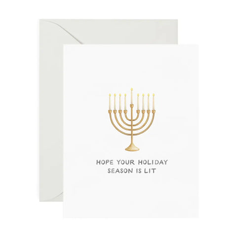 White card with drawing of gold menorah and white candles. Black text reads "hope your holiday season is lit" 