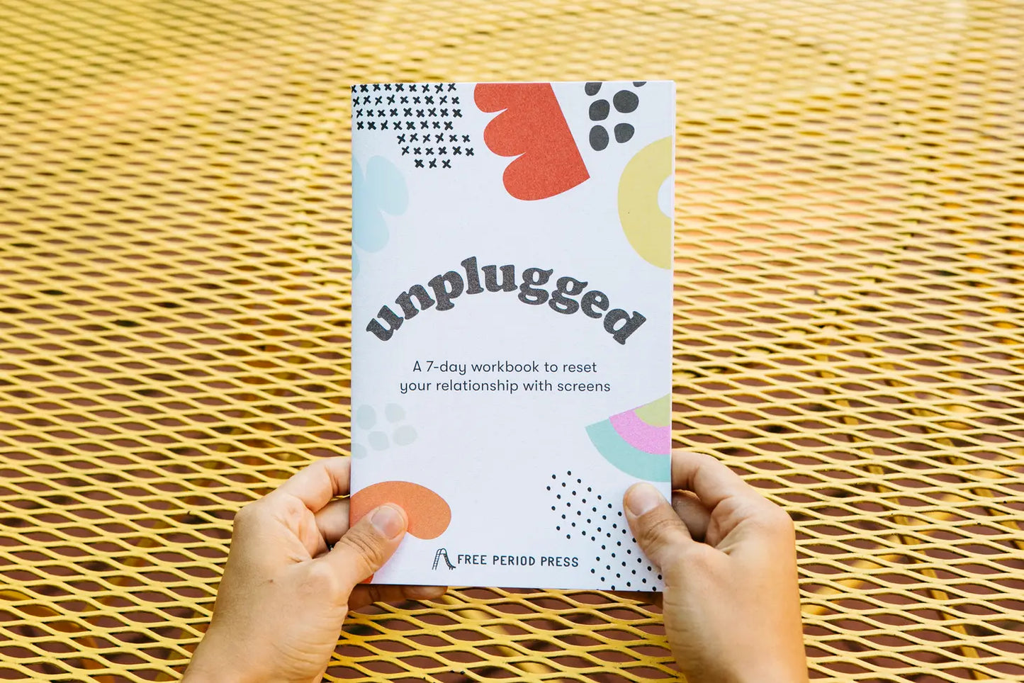 Person holding booklet. White paper with black text reading "unplugged. A 7-day workbook to reset your relationship with screens." Red, orange, yellow, and black blob illustrations. 