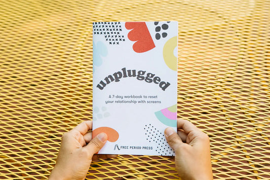 Person holding booklet. White paper with black text reading "unplugged. A 7-day workbook to reset your relationship with screens." Red, orange, yellow, and black blob illustrations. 