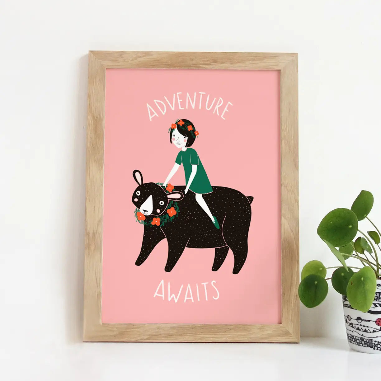 Art print with pink background and illustration of a kid on a black bear. White text reads "adventure awaits" 