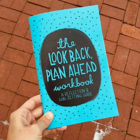 Person holding the booklet. Blue background with black polkadots. A black circle in the middle with blue text reading "the look back, plan ahead workbook. A reflection and goal setting guide"