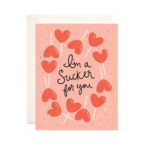 White card with a peach background. Illustrations of red heart-shaped lollipops. Black text reads "I'm a sucker for you" 