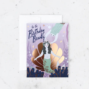 White card with a purple background and an illustration of two mermaids and a shiny gold seashell. Navy text reads "for the birthday beauty"
