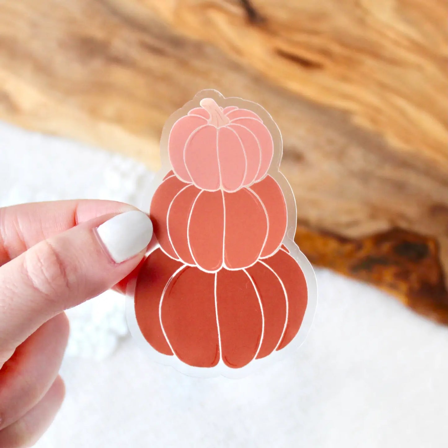 Hand holding a sticker of three pumpkins stacked on top of one another. Pumpkins are dark orange, orange, and light pink