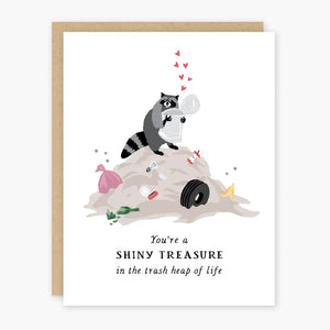 White card with raccoon holding a trash can on a trash heap. Black text reads "you're a shiny treasure in the trash heap of life"