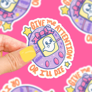 Digital pet shaped sticker. Purple and pink buttons. Orange text reads "give me attention or I'll die." White ghost on the screen of the digital pet.