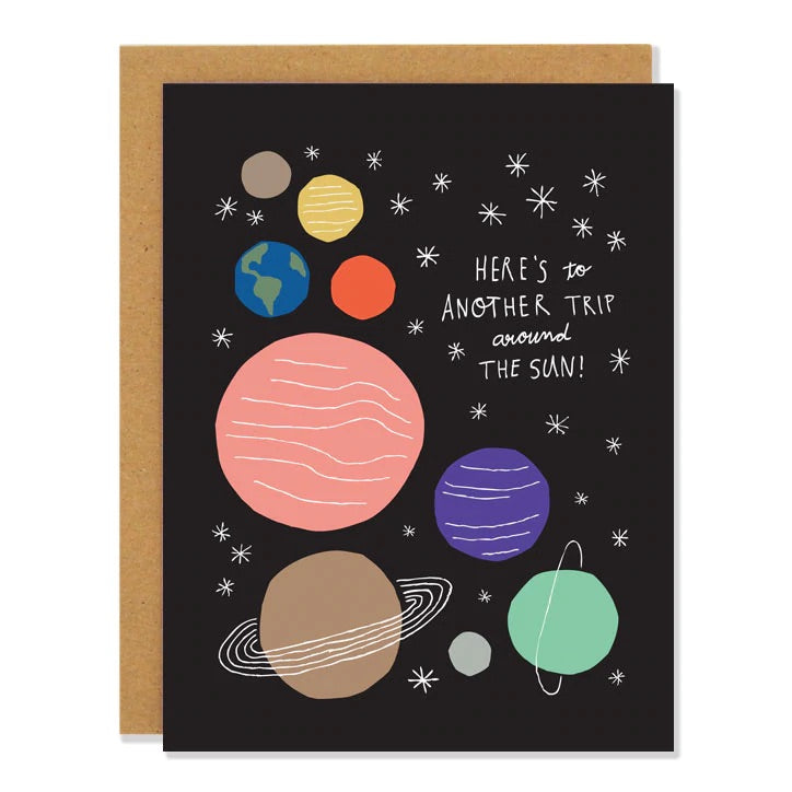 Black card with colorful planets. White text reads "here's to another trip around the sun!" Inside of card is white.