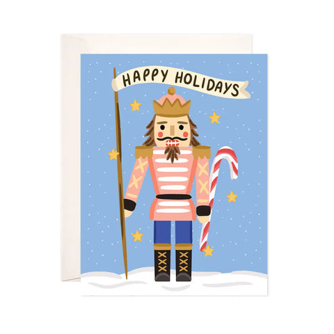 White card with a blue background and illustration of a nutcracker holding a candy cane and a flag. The flag has black text on it reading "happy holidays"
