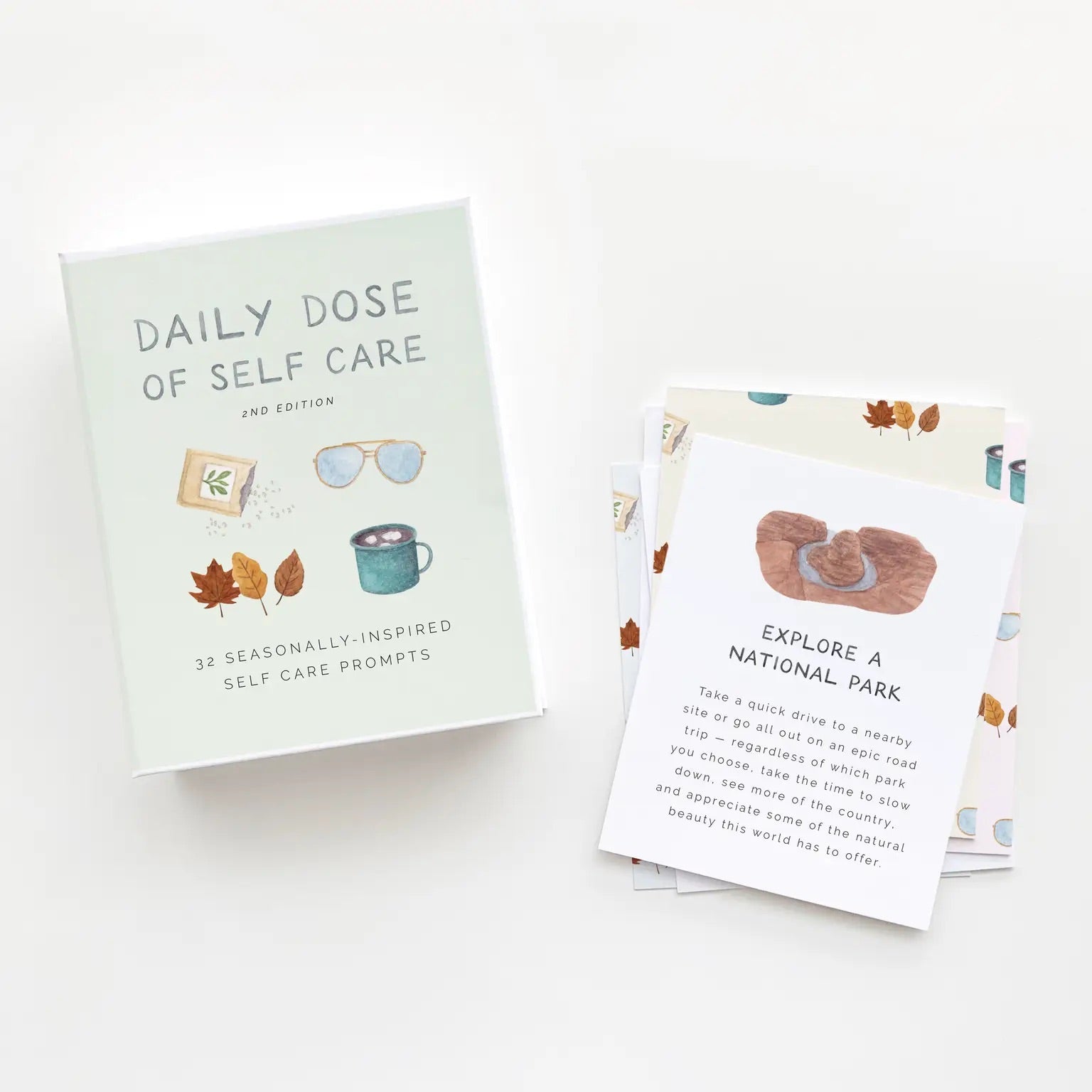 White box with black text reading "daily doses of self care." Box has drawings of leaves, seeds, sunglasses, and a mug of hot chocolate. One card is displayed with the prompt to "explore a national park." 