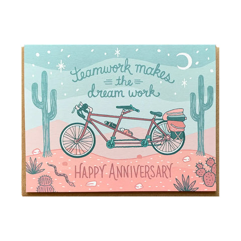 White card with light turqouise and coral background of a desert scene. A coral tandem bicycle illustration. Teal text reads "teamwork makes the dream work." Coral text reads "happy anniversary"